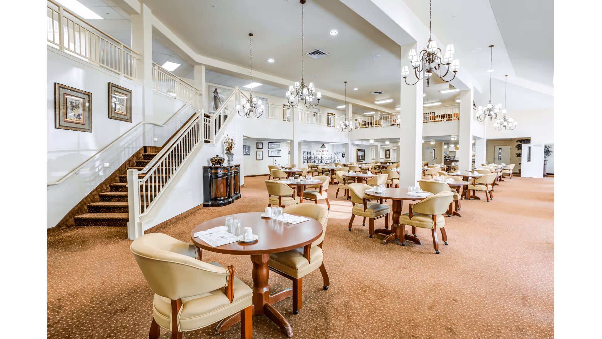 A spacious dining room with multiple round wooden tables and beige cushioned chairs arranged neatly. The room features a high ceiling with several elegant chandeliers hanging down. There is a staircase with white railings on the left side, and framed artwork is displayed on the walls. The carpet is a warm brown color, and the overall atmosphere is bright and inviting.