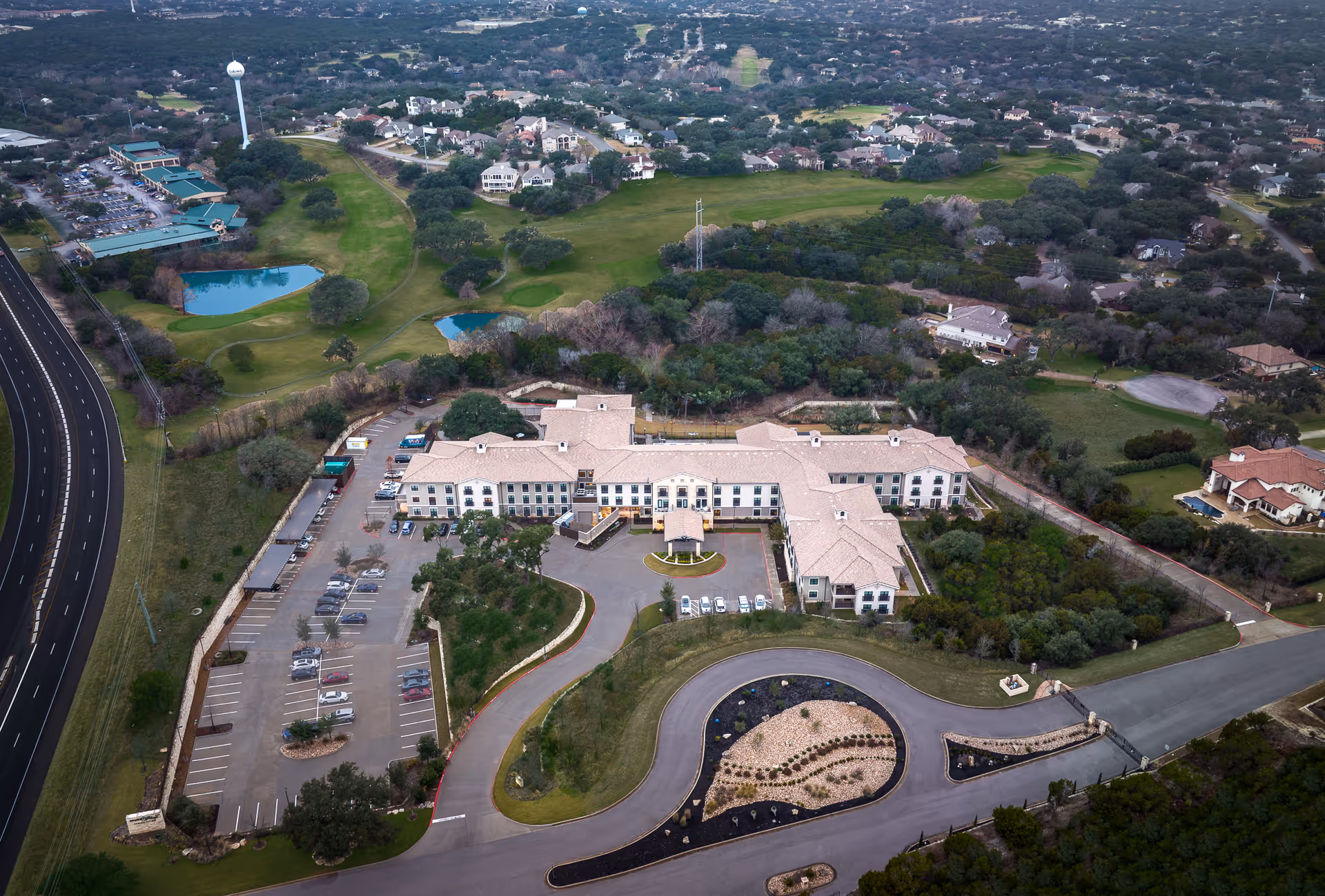 Aerial view of Belmont Village Senior Living Lakeway, showing a large, multi-wing building surrounded by trees and greenery. The facility has a circular driveway with landscaped areas and a parking lot with several cars. Nearby are residential homes, a golf course with ponds, and a water tower in the distance.