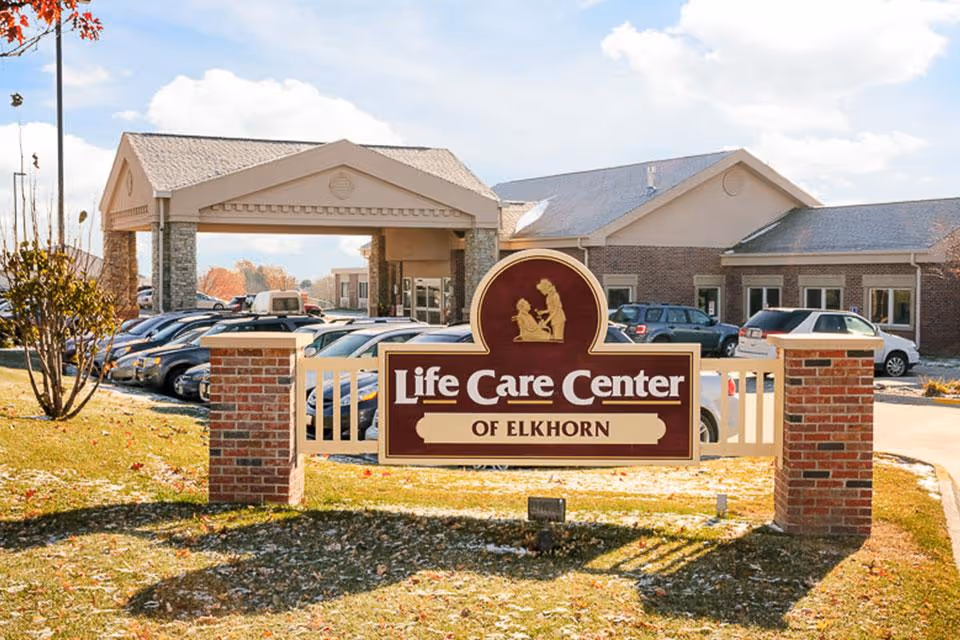 Exterior view of Life Care Center of Elkhorn showing the main entrance with a covered drop-off area, parked cars, and a large sign in the foreground displaying the facility's name.