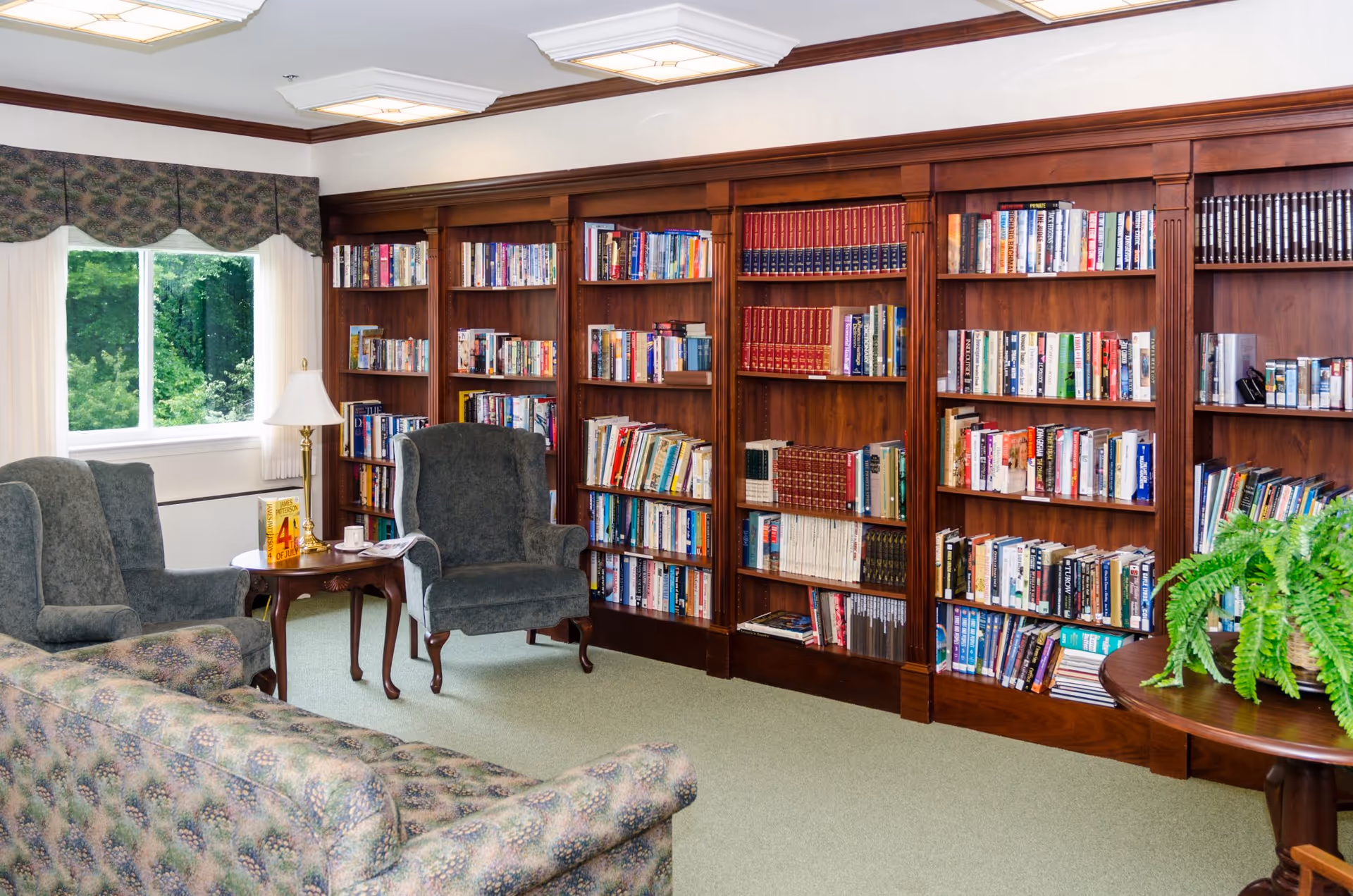 A cozy library room with wooden bookshelves filled with books, two upholstered armchairs, a patterned sofa, a wooden side table with a lamp and a book, a window with curtains showing green trees outside, and a round table with a green potted plant.