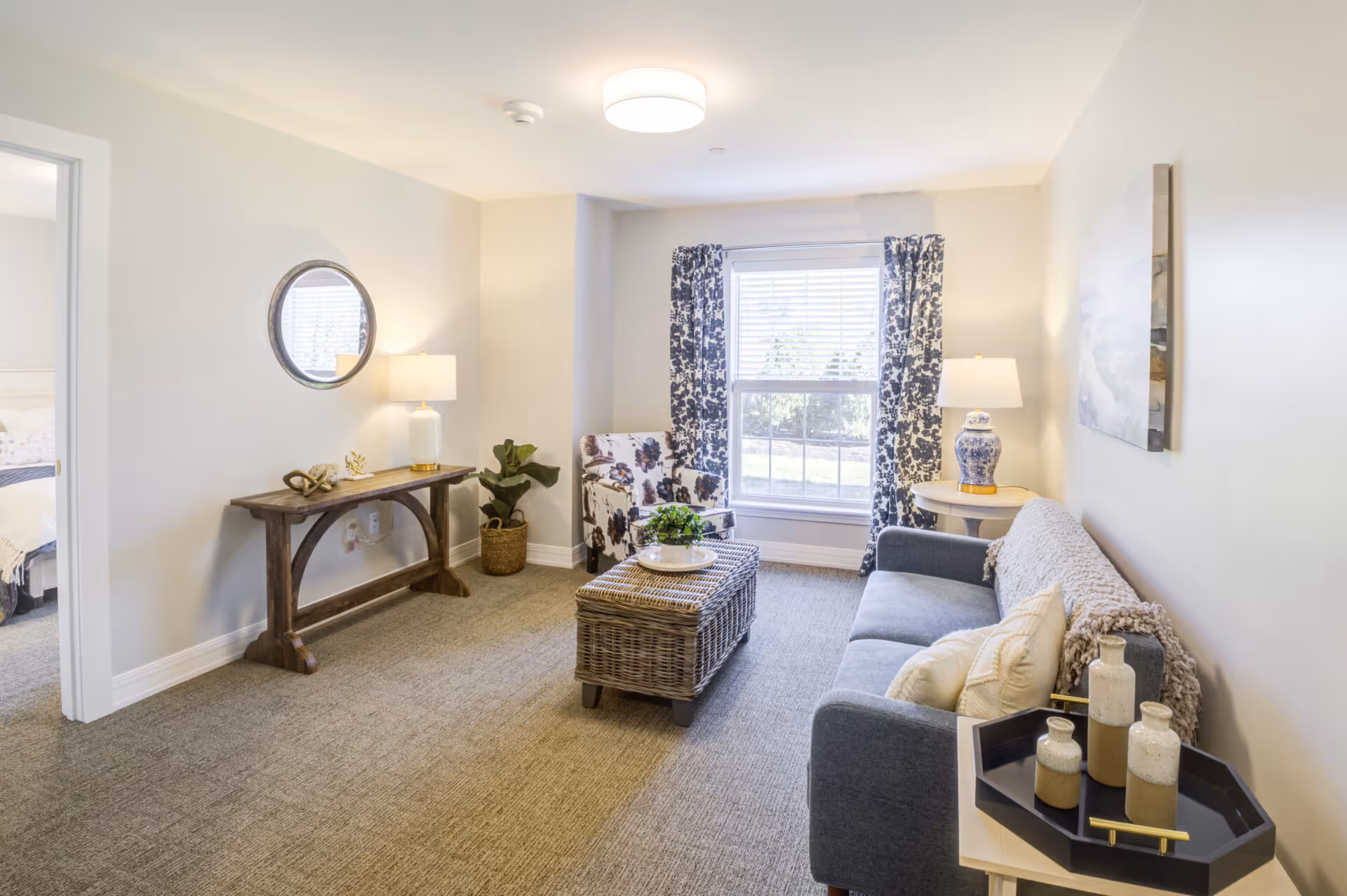 A bright and cozy living room with a gray sofa adorned with pillows and a throw blanket, a floral armchair, a wicker coffee table with a plant centerpiece, and a wooden console table with a lamp and decorative items. A large window with patterned curtains lets in natural light, and a round mirror hangs on the wall above the console table. The room has beige walls and carpeted flooring.