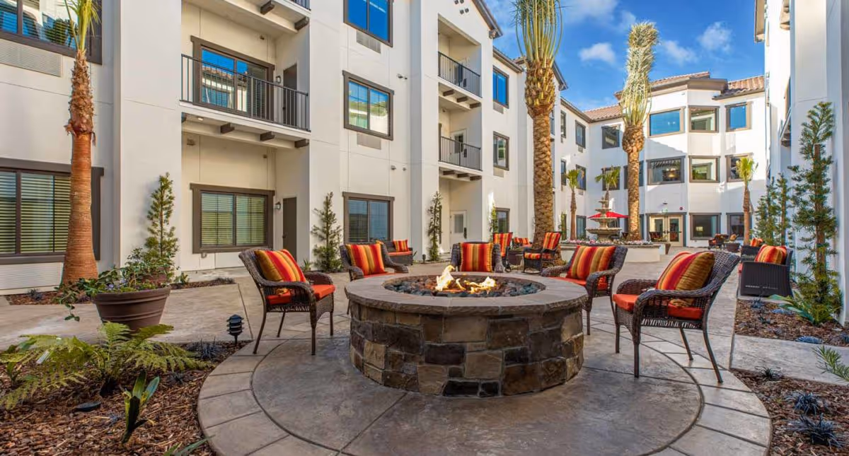 Sunlit courtyard with a central stone fire pit surrounded by wicker chairs with colorful cushions, palm trees, and a multi-story building around it.
