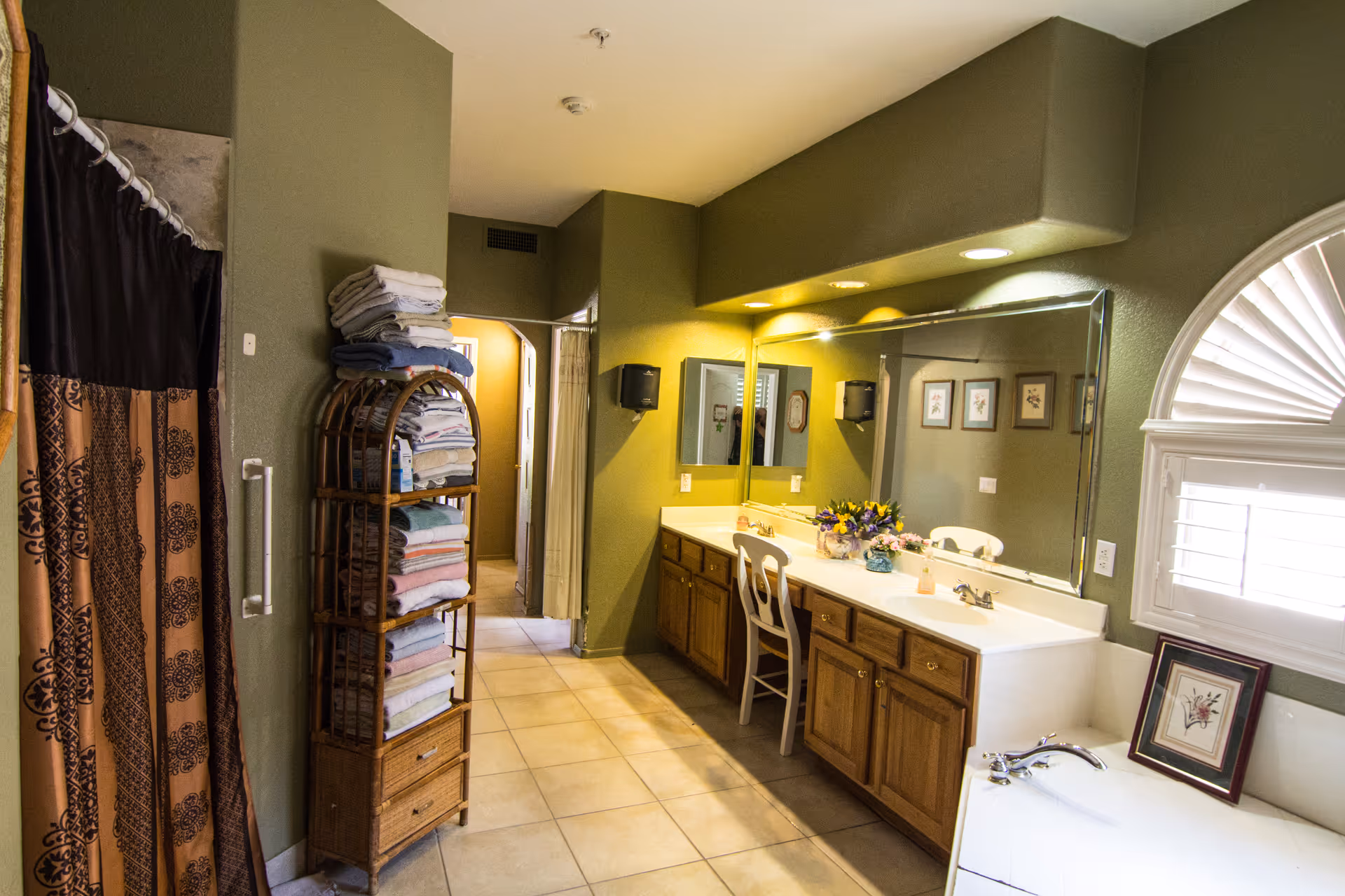A spacious bathroom with green walls, a large mirror above a double sink vanity with wooden cabinets, a chair in front of the sink, a window with white shutters, a bathtub with a framed picture on the edge, a tall rack filled with folded towels, and a shower with a patterned curtain.