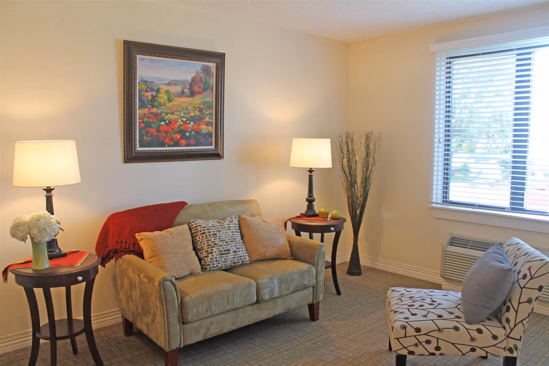 A cozy living room with a loveseat, patterned armchair, side tables with lamps, wall art, and a window with blinds.
