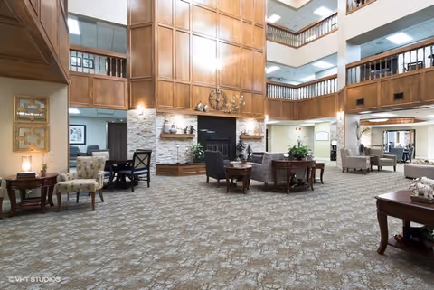 Spacious and well-lit common area in a senior living facility with multiple seating arrangements including armchairs and small tables. The room features a large wooden paneled fireplace with stone accents, high ceilings, and balconies overlooking the space from the upper floors. The carpet has a patterned design and the walls are decorated with framed art and mirrors.