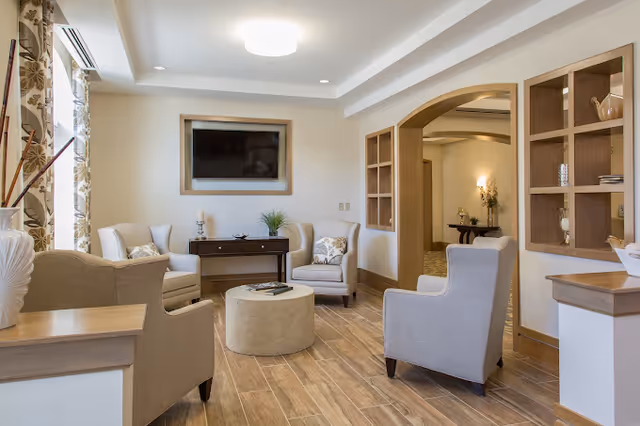 A cozy living room area with four beige armchairs arranged around a round beige ottoman. There is a wooden console table against the wall with decorative items and a flat-screen TV mounted above it. The room features wood-look tile flooring, built-in wooden shelves, and an arched doorway leading to another space. Natural light comes in through a window with floral curtains on the left.