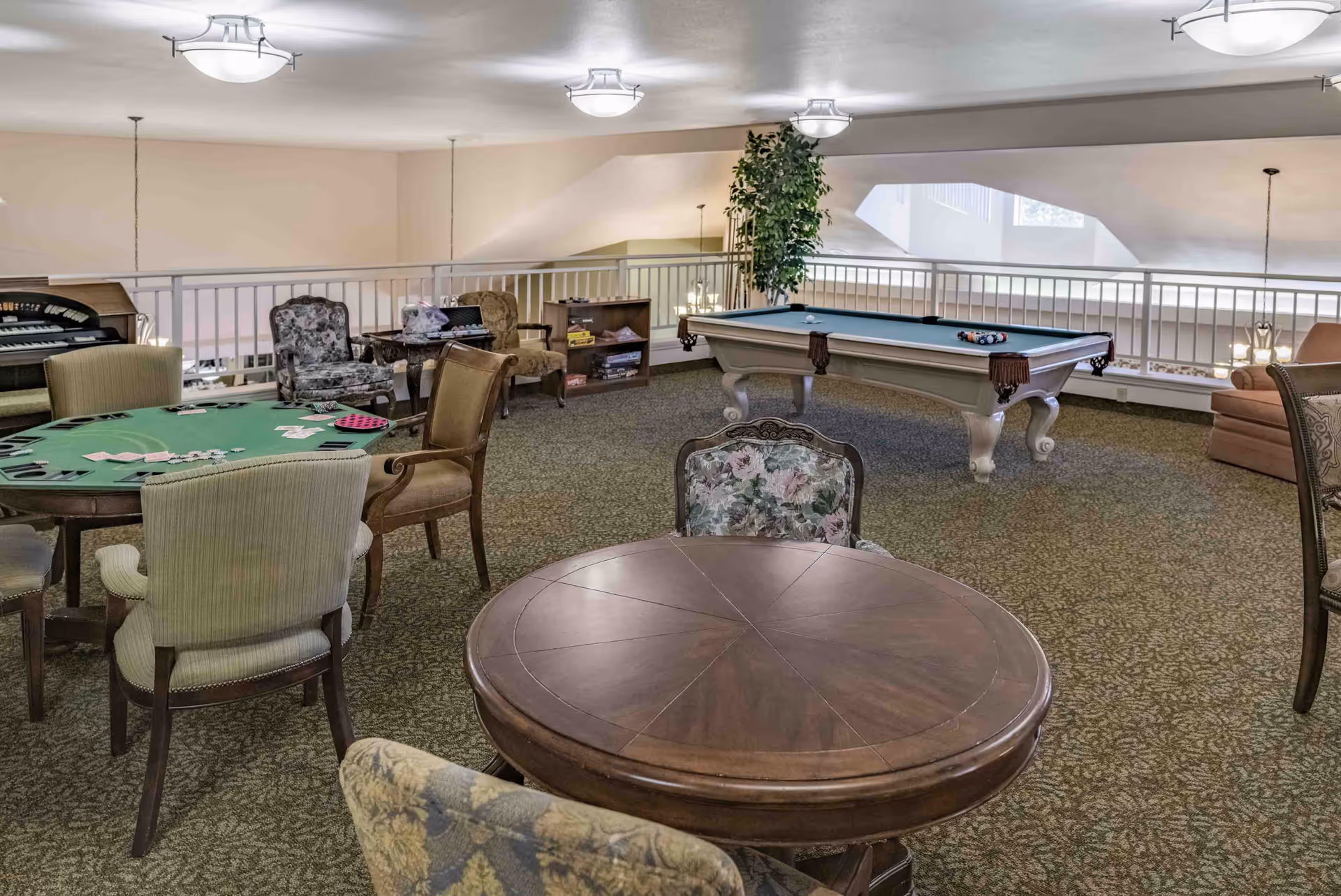 A spacious recreational room with a pool table, a card table with playing cards and poker chips, several upholstered chairs, a small bookshelf with board games, and an organ in the corner. The room has carpeted flooring, soft lighting from ceiling fixtures, and a railing overlooking a lower level.
