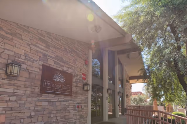 Exterior view of Heritage Court Post Acute Of Scottsdale showing a stone facade with the facility's sign mounted on the wall, large windows, outdoor lighting fixtures, and a shaded walkway with trees nearby.