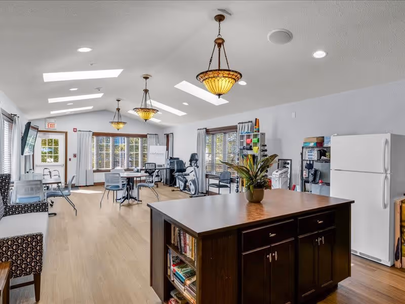 A bright, spacious common area with wooden flooring and multiple skylights. The room features a large dark wooden island with bookshelves and a potted plant on top, several chairs and tables, exercise equipment, a white refrigerator, and large windows with white curtains letting in natural light. Three decorative pendant lights hang from the ceiling.