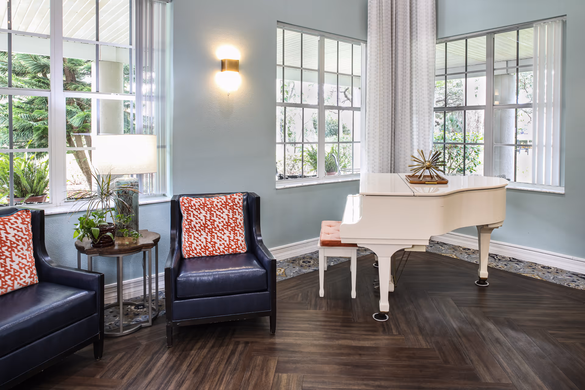 A bright and airy room with large windows showing greenery outside. Inside, there are two dark blue armchairs with red and white patterned cushions, a small round wooden side table with a lamp and plants, and a white grand piano with a matching white bench. The floor has dark wood herringbone pattern and the walls are painted light blue.