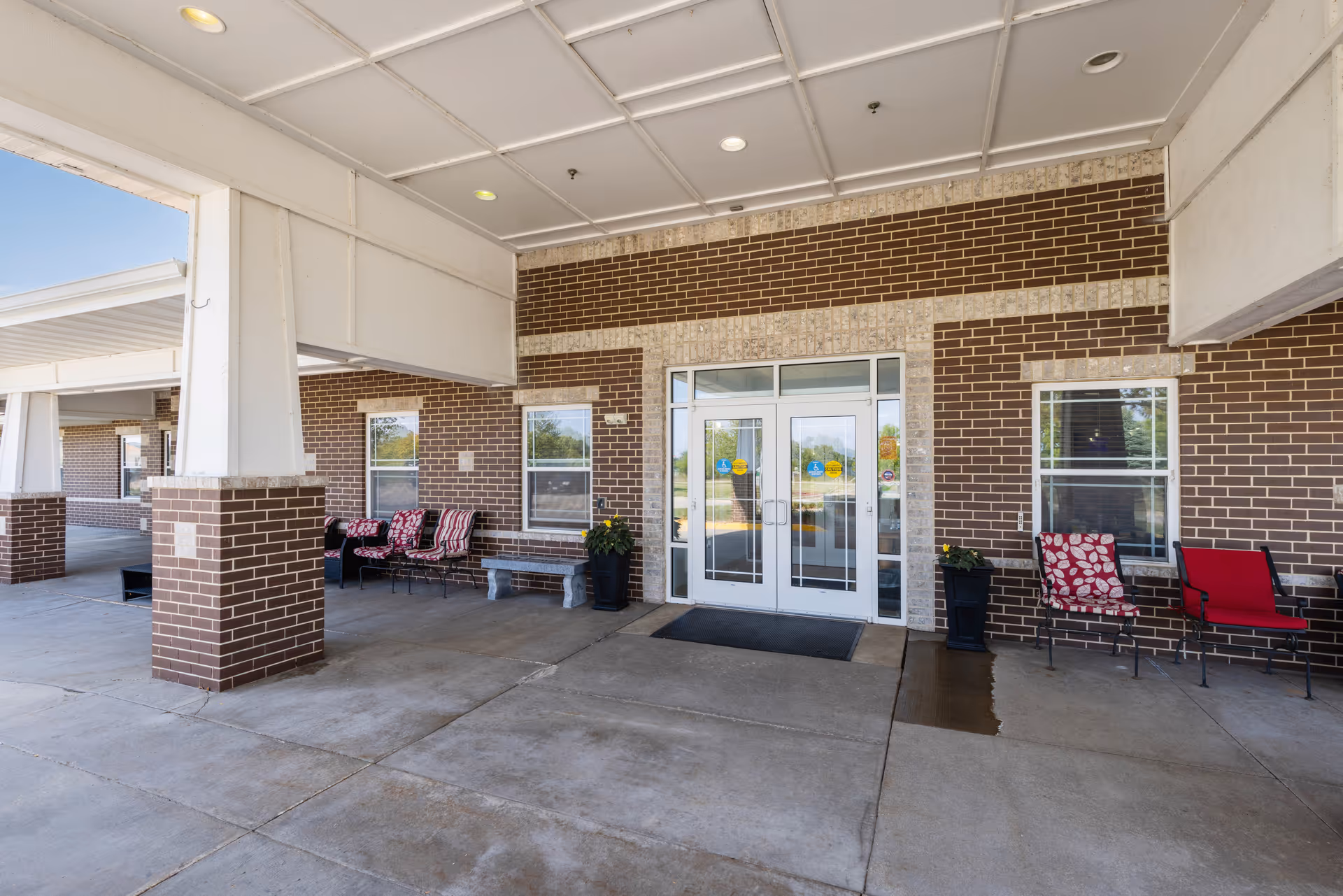 Covered building entrance with double glass doors, brick facade and outdoor seating.