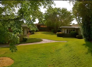 A well-maintained outdoor garden area with a winding paved pathway surrounded by green grass and trees, with single-story buildings visible in the background.