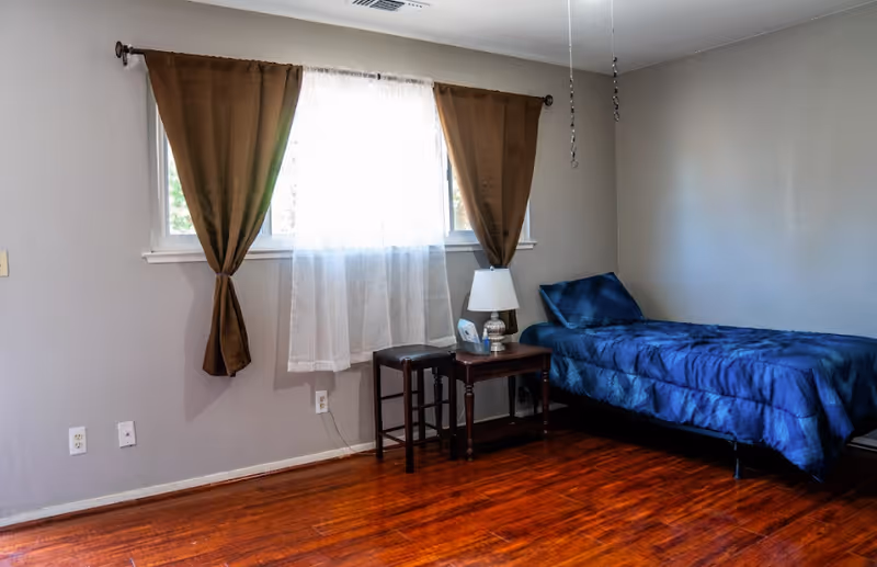 A simple bedroom with a single bed covered in a blue comforter, a small wooden side table with a lamp and a tissue box, two stools, and a window with brown curtains and a sheer white curtain in the center. The room has wooden flooring and light gray walls.