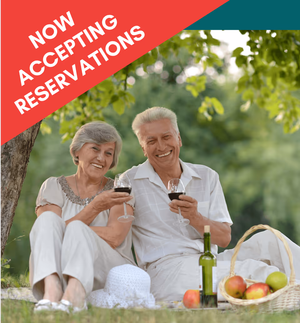 An elderly couple sitting on the grass outdoors having a picnic, smiling and holding glasses of red wine. There is a basket with apples and a bottle of wine beside them. Green trees and foliage are in the background.