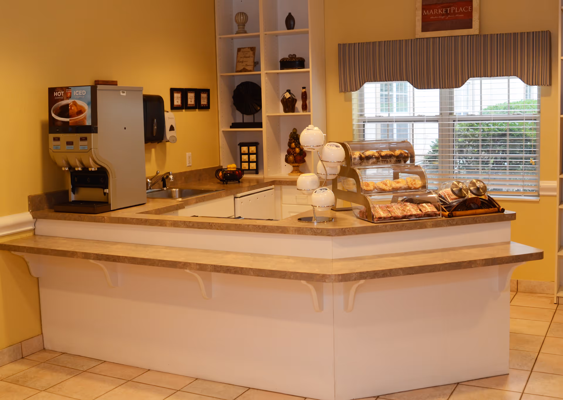 A small food service counter area with a coffee and iced beverage dispenser on the left, a sink, and a display case containing pastries and snacks. Behind the counter are shelves with decorative items and a window with blinds and a striped valance. The walls are painted yellow and the floor is tiled.