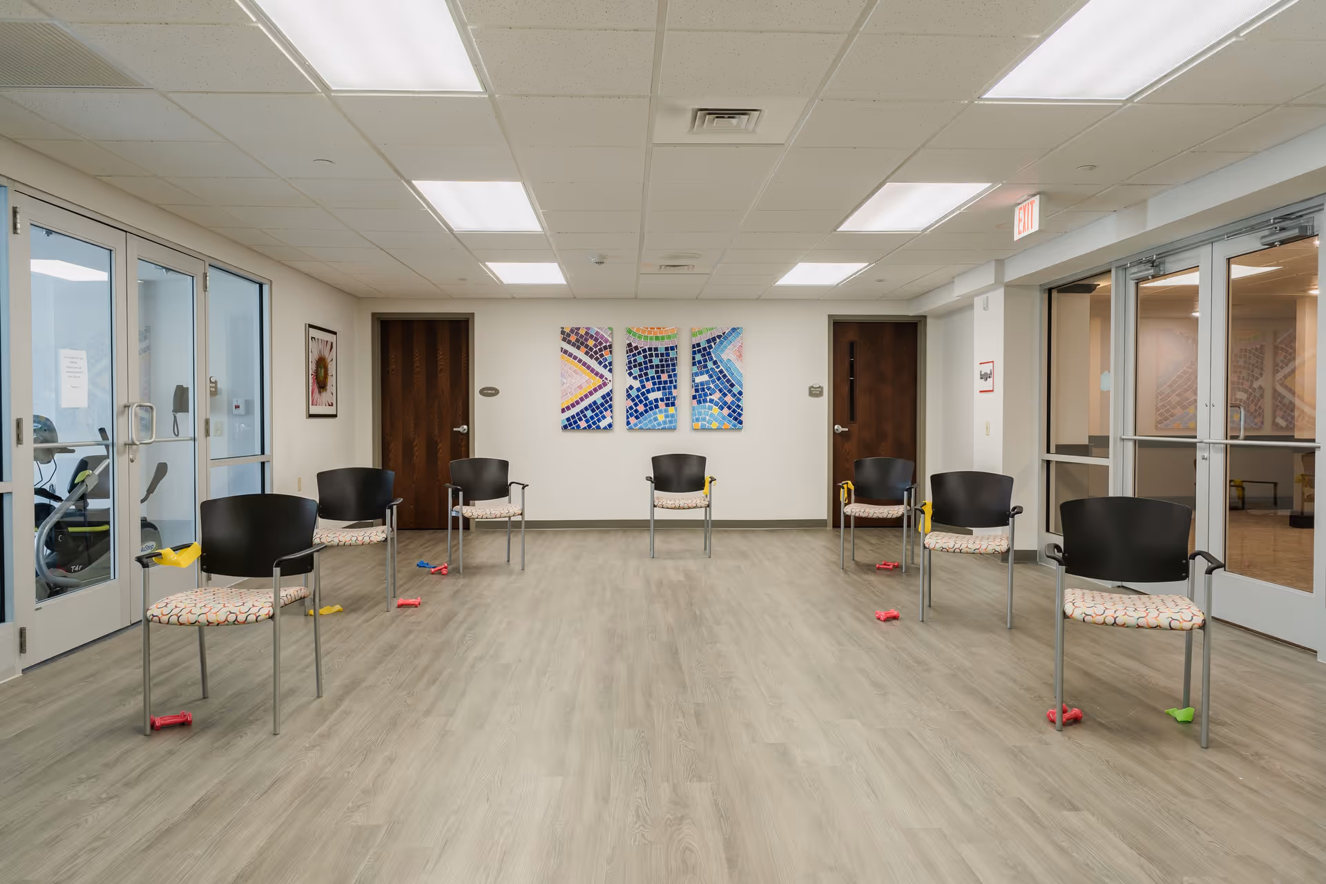 A spacious room with eight chairs arranged in two rows facing each other. Each chair has a patterned cushion and some have small exercise weights and resistance bands nearby. The room has light wood flooring, white walls, and a drop ceiling with fluorescent lighting. There are two wooden doors on the far wall with colorful mosaic artwork between them. Glass doors on both sides lead to other rooms, one of which contains exercise equipment.