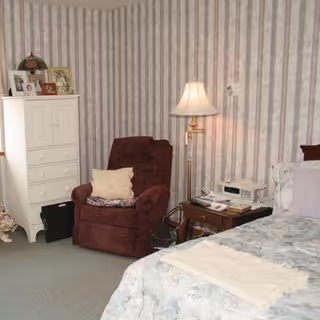 A cozy bedroom with a floral-patterned bedspread on a bed, a burgundy recliner chair with a pillow, a wooden nightstand with a lamp, telephone, and books, and a white dresser with framed photos and decorative items. The walls have striped wallpaper.