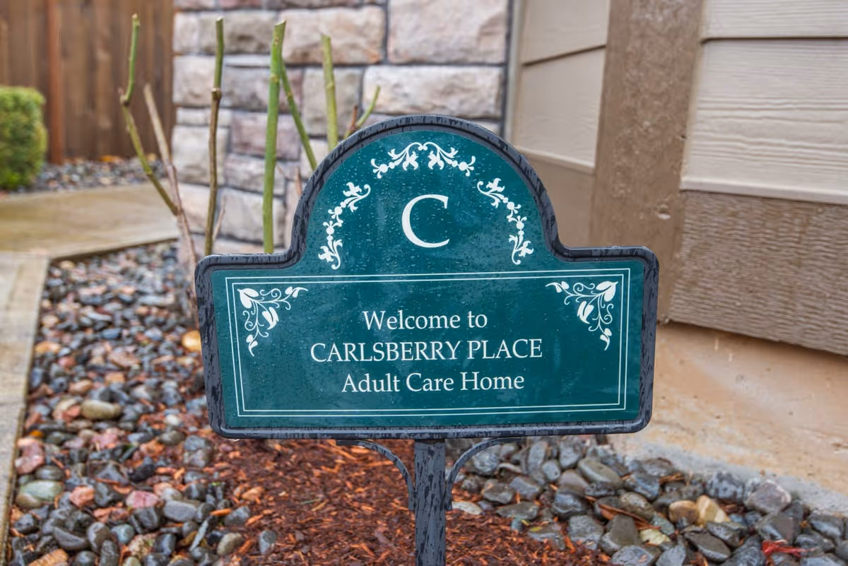 A green decorative sign with white floral designs that reads 'Welcome to CARLSBERRY PLACE Adult Care Home' placed outdoors in a landscaped area with rocks and mulch near a building wall.
