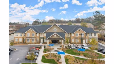 Front exterior view of a large two-story senior living facility building with a covered entrance, surrounded by landscaped gardens, walkways, parked cars, and blue sky with scattered clouds.