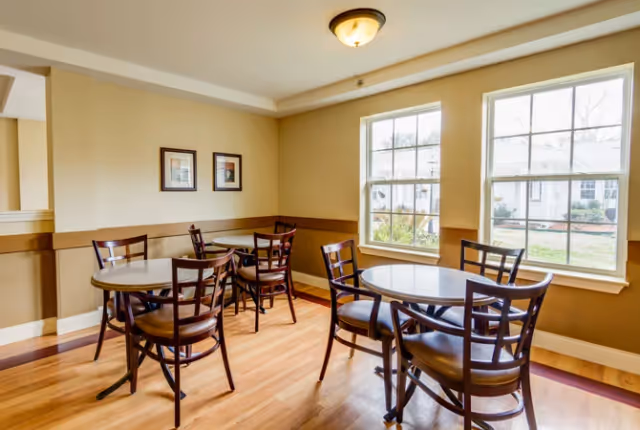 A bright dining area with wooden floors, beige walls, and two large windows letting in natural light. The room contains three round tables, each surrounded by four wooden chairs with cushioned seats. Two framed pictures hang on the wall.