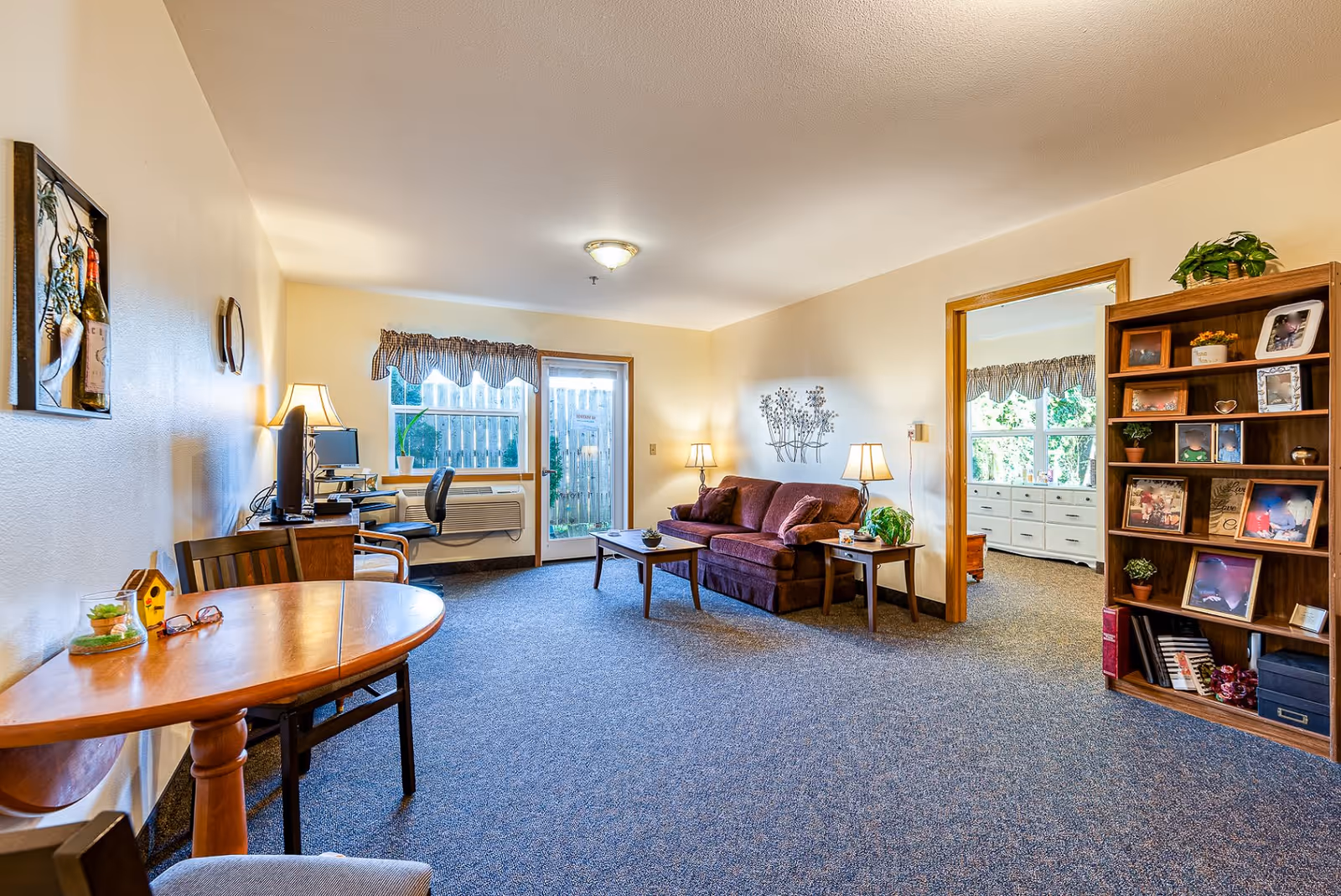 A cozy living room in an assisted living facility featuring a purple sofa with pillows, a wooden coffee table, two side tables with lamps, a wooden bookshelf filled with framed photos and plants, a small round dining table with chairs, a desk with a computer and chair near a window, and a door leading outside. The room has beige walls, blue carpet, and a window with a valance.