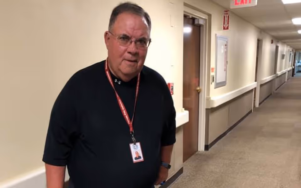 An older man wearing glasses and a black shirt stands in a hallway of a care facility. He has a red lanyard with an ID badge around his neck. The hallway has beige walls, a carpeted floor, and several doors along the side. An exit sign and fire safety equipment are visible on the wall.