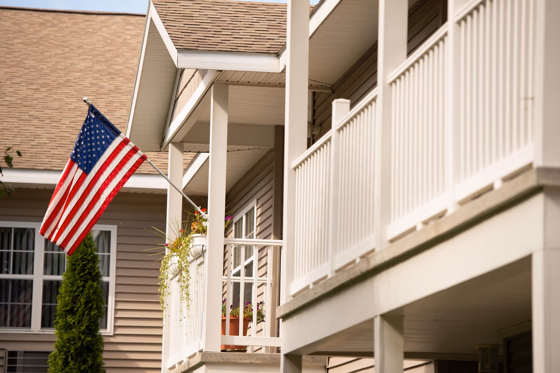 Close-up view of a beige building exterior with white railings on a balcony. An American flag is mounted on a pole attached to the balcony, and there are flower pots with green plants and flowers on the railing. A window and a tall green shrub are visible in the background.