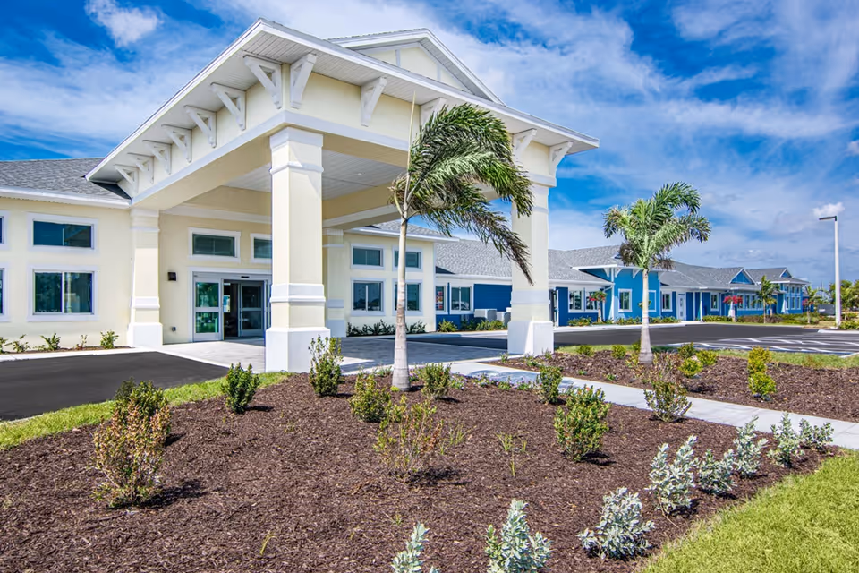 Front entrance of a light-colored senior living building with a covered porte-cochère, palm trees, and landscaped beds under a blue sky.