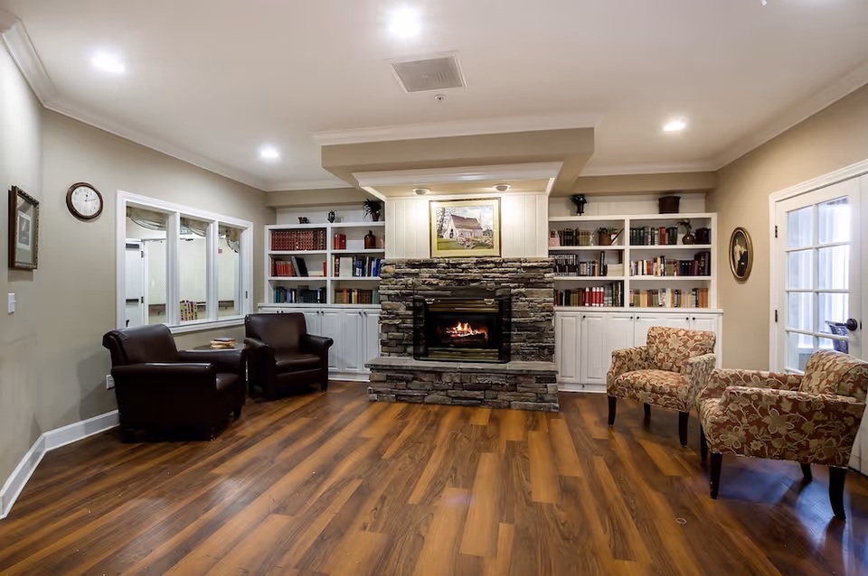 A cozy living room area with a stone fireplace in the center, flanked by built-in white bookshelves filled with books and decorative items. There are two dark brown leather armchairs on the left side and two floral-patterned armchairs on the right side. The room has wooden flooring, beige walls, recessed ceiling lights, a wall clock, and a framed picture above the fireplace.