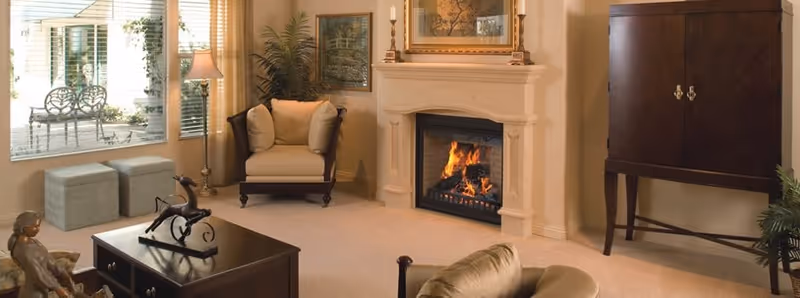 A cozy living room with a lit fireplace, beige carpet, and neutral-colored walls. The room features a cushioned armchair, a wooden coffee table with decorative items, a floor lamp, and a large window with a view of an outdoor patio area with chairs. There is also a dark wooden cabinet and framed artwork above the fireplace.