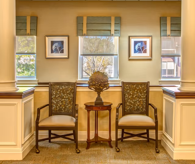 Two wooden armchairs with patterned cushions placed on either side of a small round wooden table with a decorative flower sculpture on top, set against a wall with three windows and two framed botanical prints in a well-lit room.
