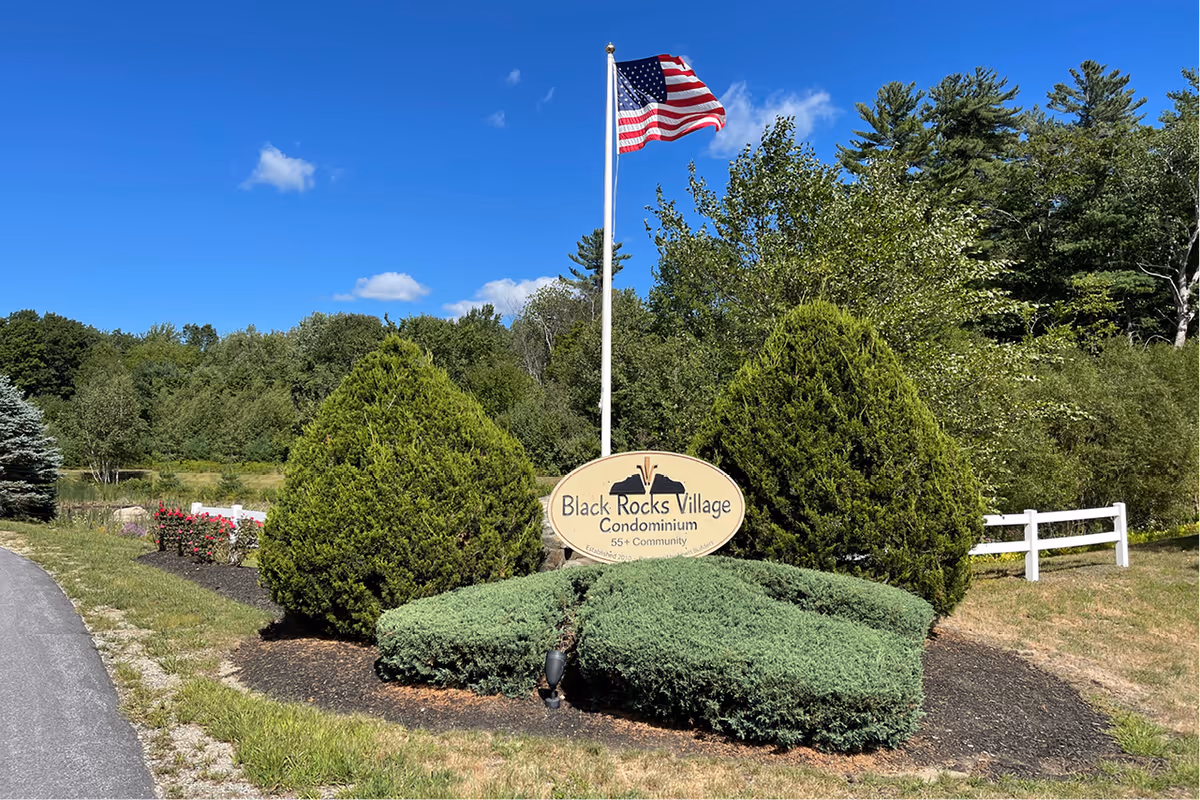 Outdoor view of the entrance sign for Black Rocks Village Condominium, a 55+ community, surrounded by green bushes and trees under a clear blue sky with an American flag flying on a flagpole above the sign.