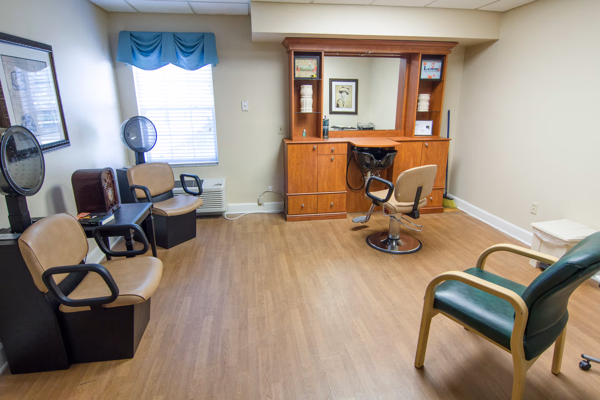 A small salon room with a wooden floor, featuring a hair washing station with a chair and sink, two hair drying chairs with hooded dryers, a green cushioned chair, a window with blue valance, framed artwork on the walls, and a wooden cabinet with a large mirror.