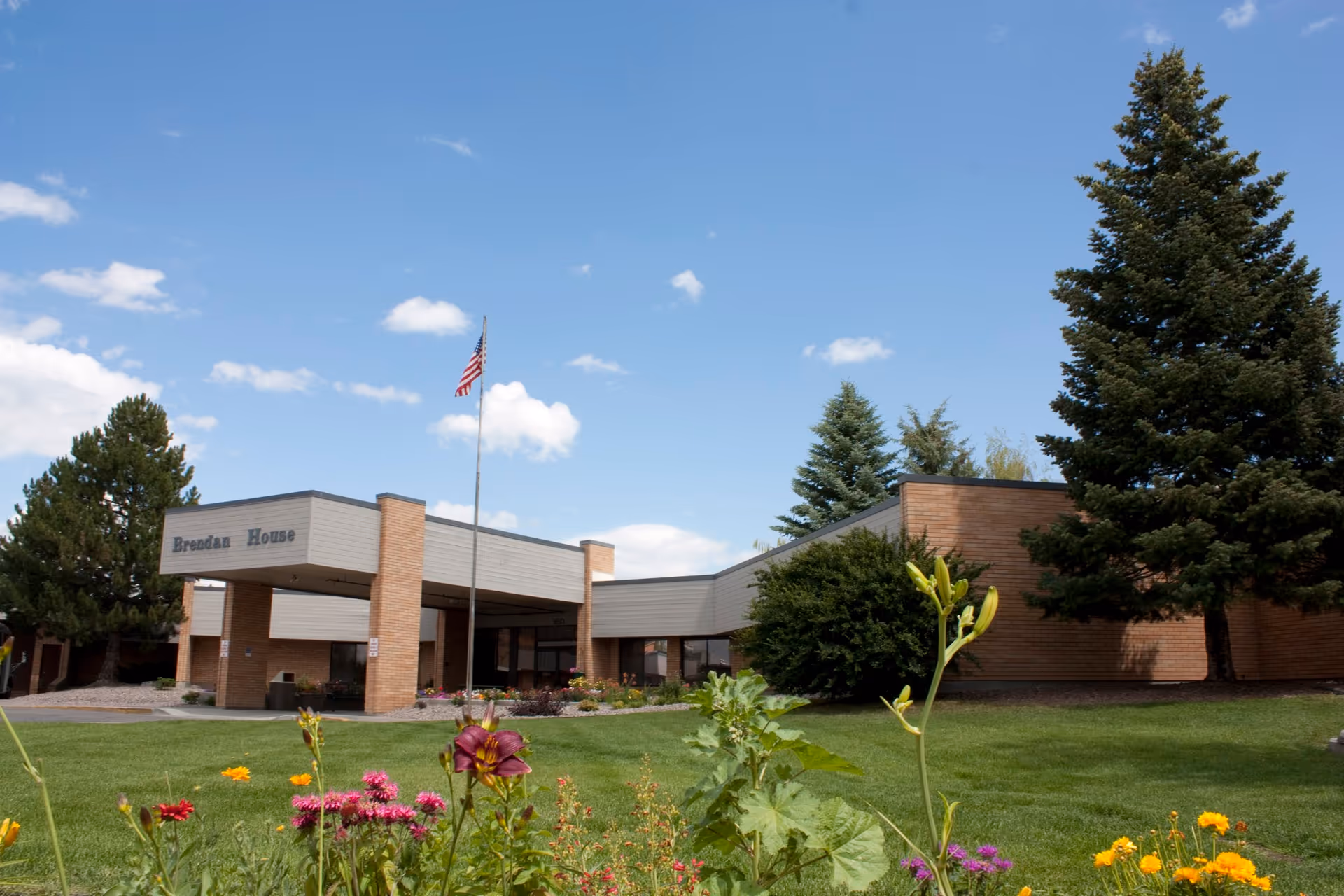 Exterior view of Logan Health Brendan House building with a covered entrance, an American flag on a flagpole, green lawn, colorful flowers, and tall evergreen trees under a blue sky with scattered clouds.