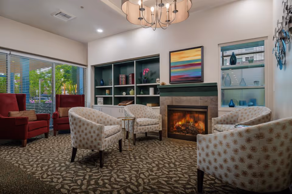 Cozy common lounge with patterned armchairs arranged around a lit fireplace, built-in shelves, and large windows.
