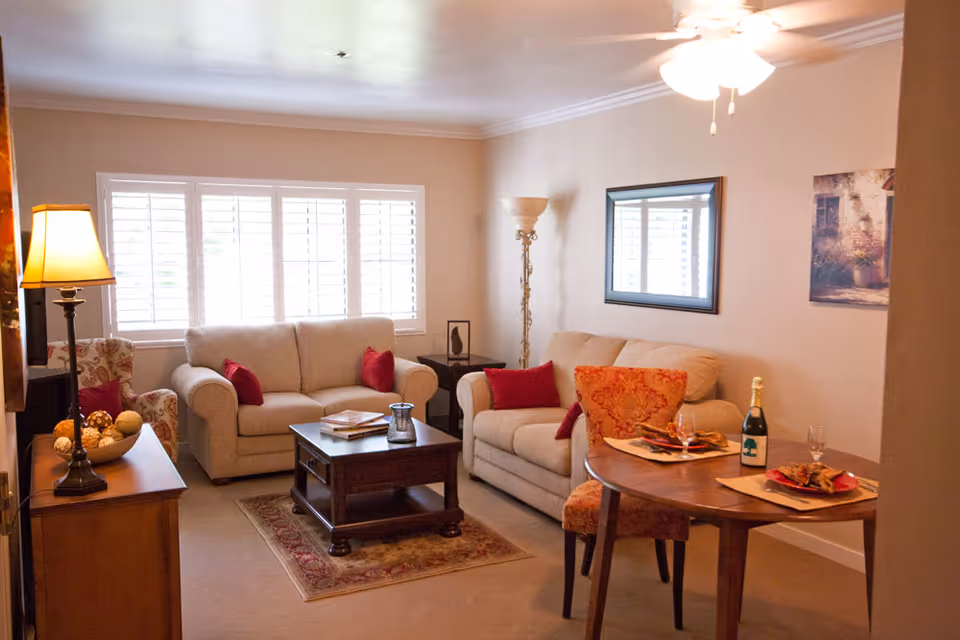 Comfortable living room with two beige sofas, a central coffee table, a small dining table set for two, lamps, and a large shuttered window.