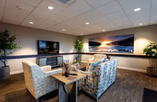 Cozy communal lounge with armchairs, a patterned sofa, TV console, plants, and a large landscape wall print.