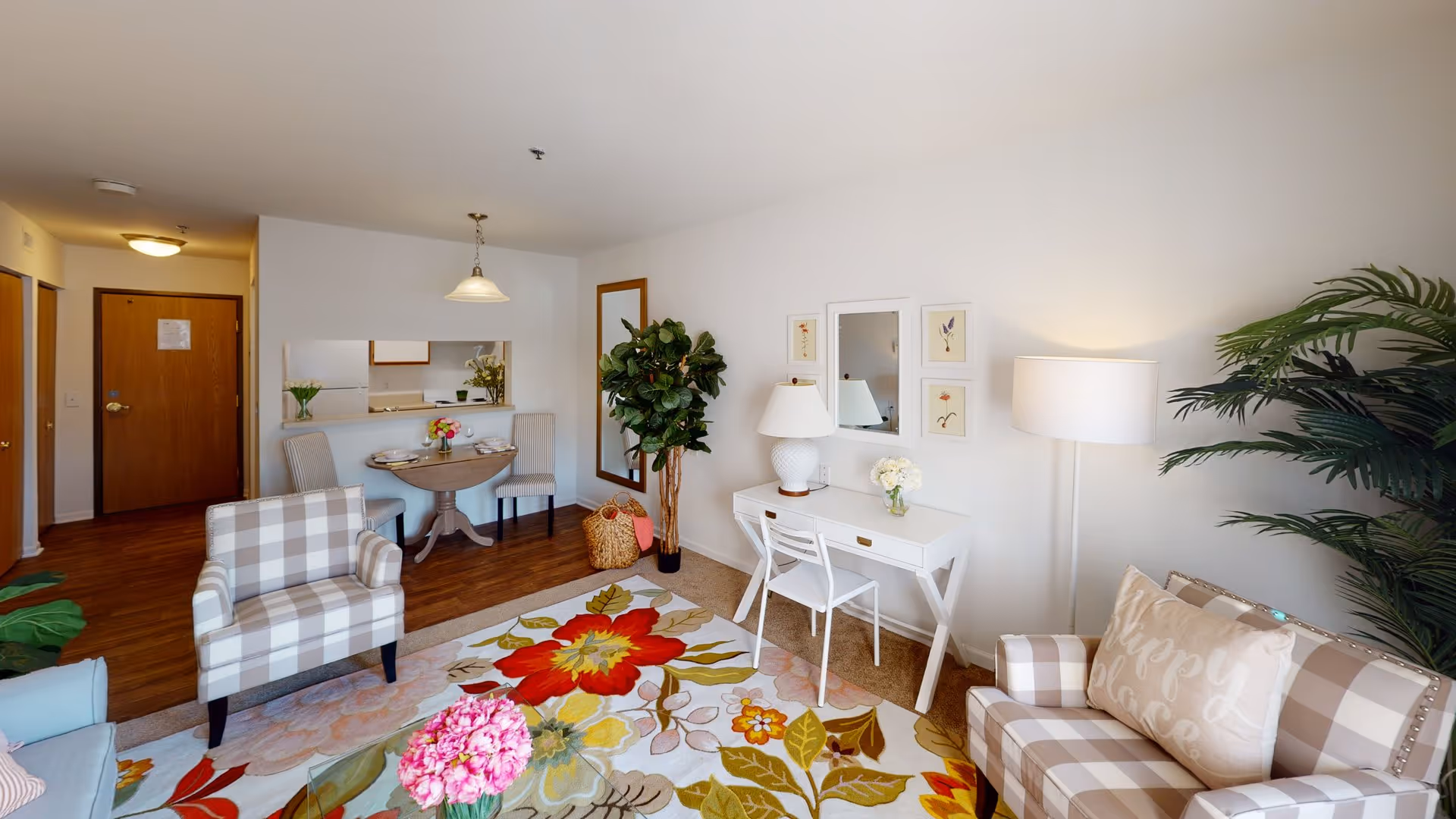 A bright and cozy living area in a senior living facility featuring a floral area rug, two plaid armchairs with a decorative pillow, a white desk with a lamp and chair, a tall floor lamp, a large potted plant, and a small dining table with two chairs near the kitchen pass-through window. The walls are white, and the flooring is a mix of wood and carpet.