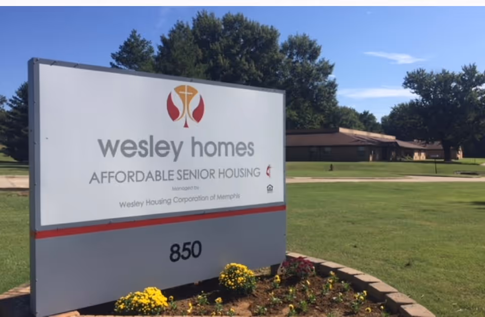 A large sign reading "wesley homes AFFORDABLE SENIOR HOUSING" with the number 850, planted flowers, and a single-story building and lawn in the background.