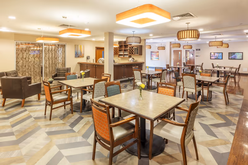 A spacious dining room with multiple square tables and chairs arranged neatly. The room features modern light fixtures hanging from the ceiling, a patterned floor, and a counter area with wine racks and cabinets in the background. There are also two armchairs near a window with patterned curtains.