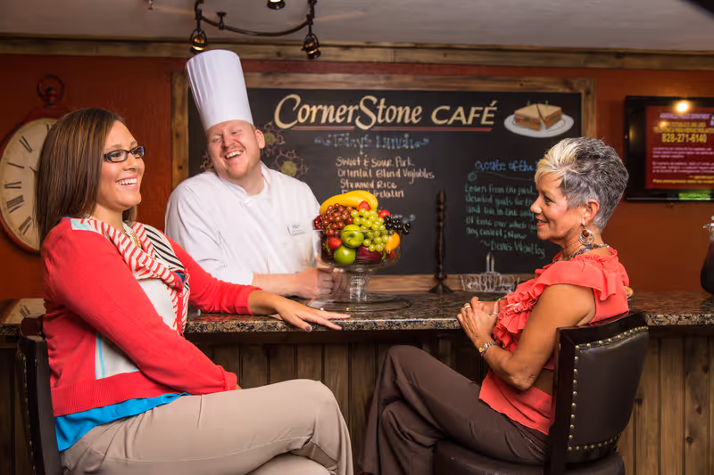 Two women sitting and smiling at a counter in a café setting, with a chef standing behind the counter smiling. A large bowl of fruit is on the counter, and a chalkboard behind them reads 'CornerStone Café' with menu items listed.