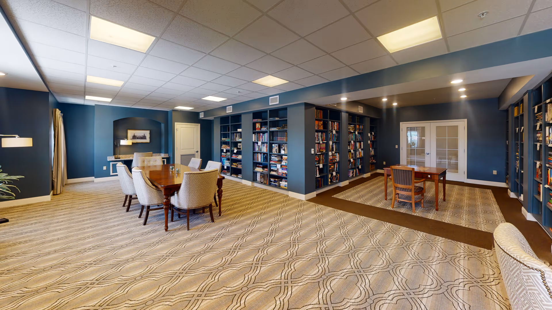 A spacious interior room with blue walls and patterned carpet featuring multiple wooden tables and upholstered chairs. The room includes built-in bookshelves filled with books along the walls, recessed lighting, and a ceiling with fluorescent lights. There are double glass doors at the back and a small desk area with a lamp and framed picture on the left side.