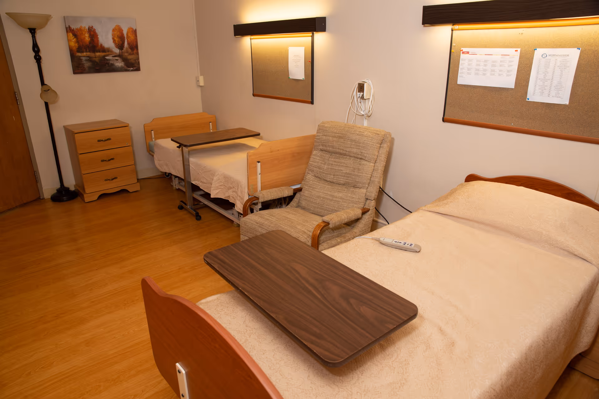 A room with two single beds, each with a beige bedspread. One bed has a remote control on it. Between the beds is a beige upholstered armchair with wooden armrests. Each bed has a wooden overbed table. There is a wooden three-drawer dresser near the door, a floor lamp, and two bulletin boards on the wall with papers pinned to them. The floor is wooden, and the walls are light-colored.