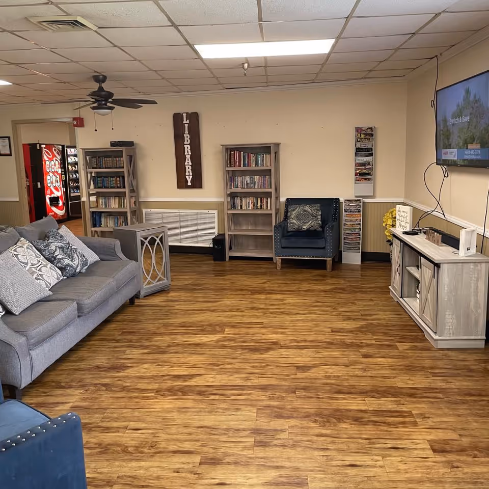 A cozy common area with wooden flooring, a gray couch with patterned pillows, a blue armchair, two wooden bookshelves filled with books, a wooden side table, and a TV mounted on the wall. A sign on the wall reads 'LIBRARY'. There is a vending machine visible in an adjacent room.
