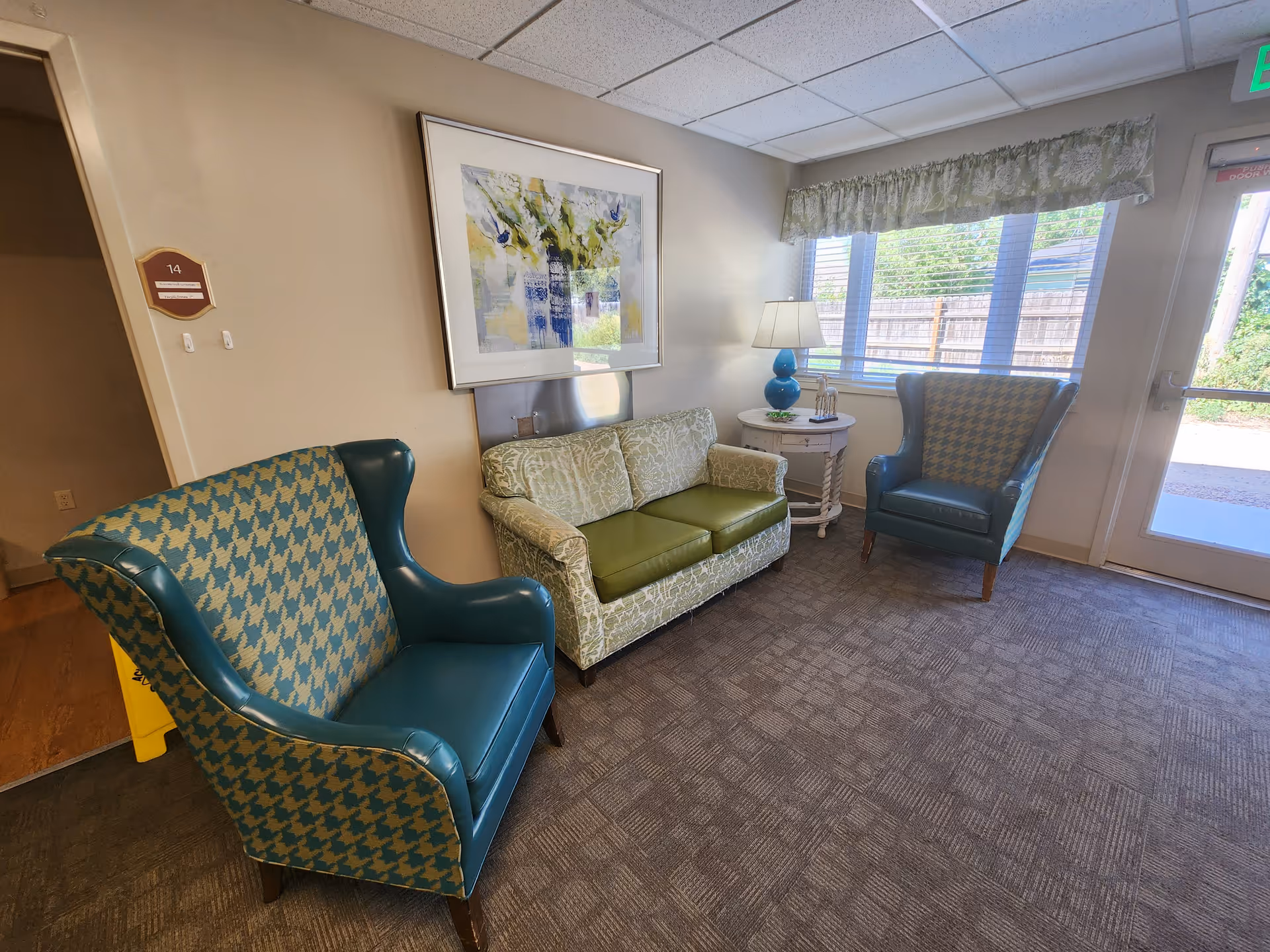 A small seating area in a senior living facility with two blue patterned armchairs and a green patterned loveseat. A round side table with a blue lamp and decorative items sits between the loveseat and one armchair. A large window with blinds and a floral valance lets in natural light. A framed abstract painting hangs on the wall above the loveseat. The carpeted floor and beige walls create a cozy atmosphere.