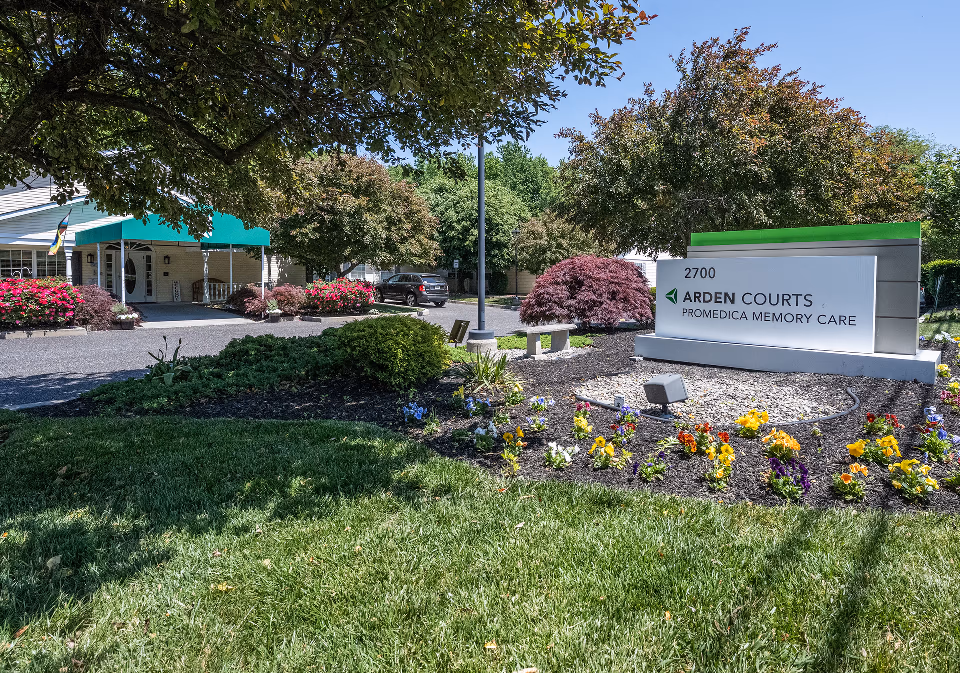 Exterior view of Arden Courts - ProMedica Memory Care Community in Cherry Hill, showing the facility's entrance with a green awning, a parking area with a vehicle, landscaped flower beds with colorful flowers, trees, and a large sign displaying the facility's name and address.