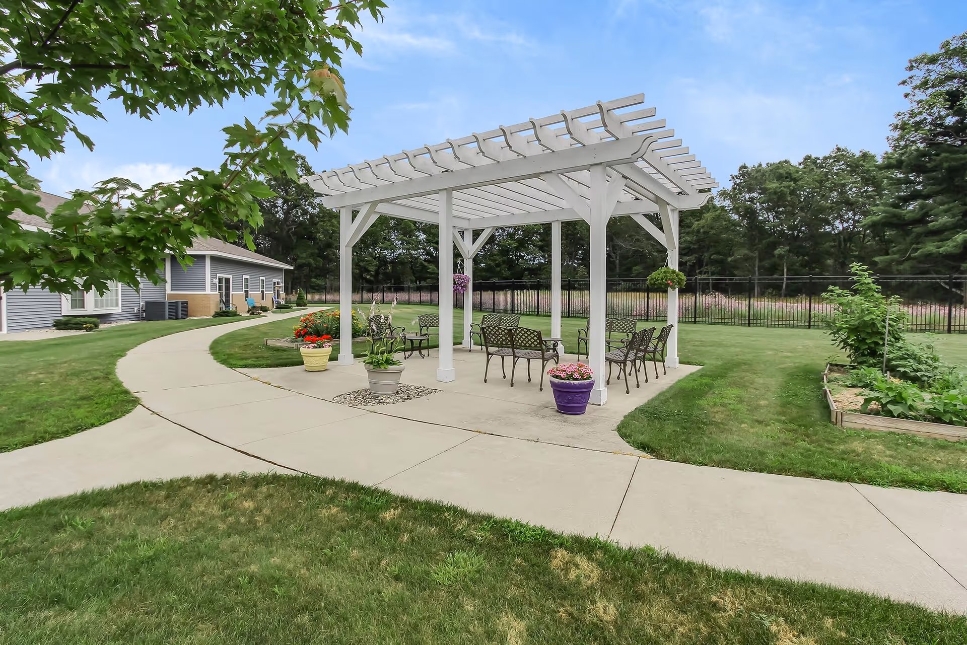 Paved courtyard with a white pergola and outdoor seating surrounded by lawn, potted plants, and a nearby building.