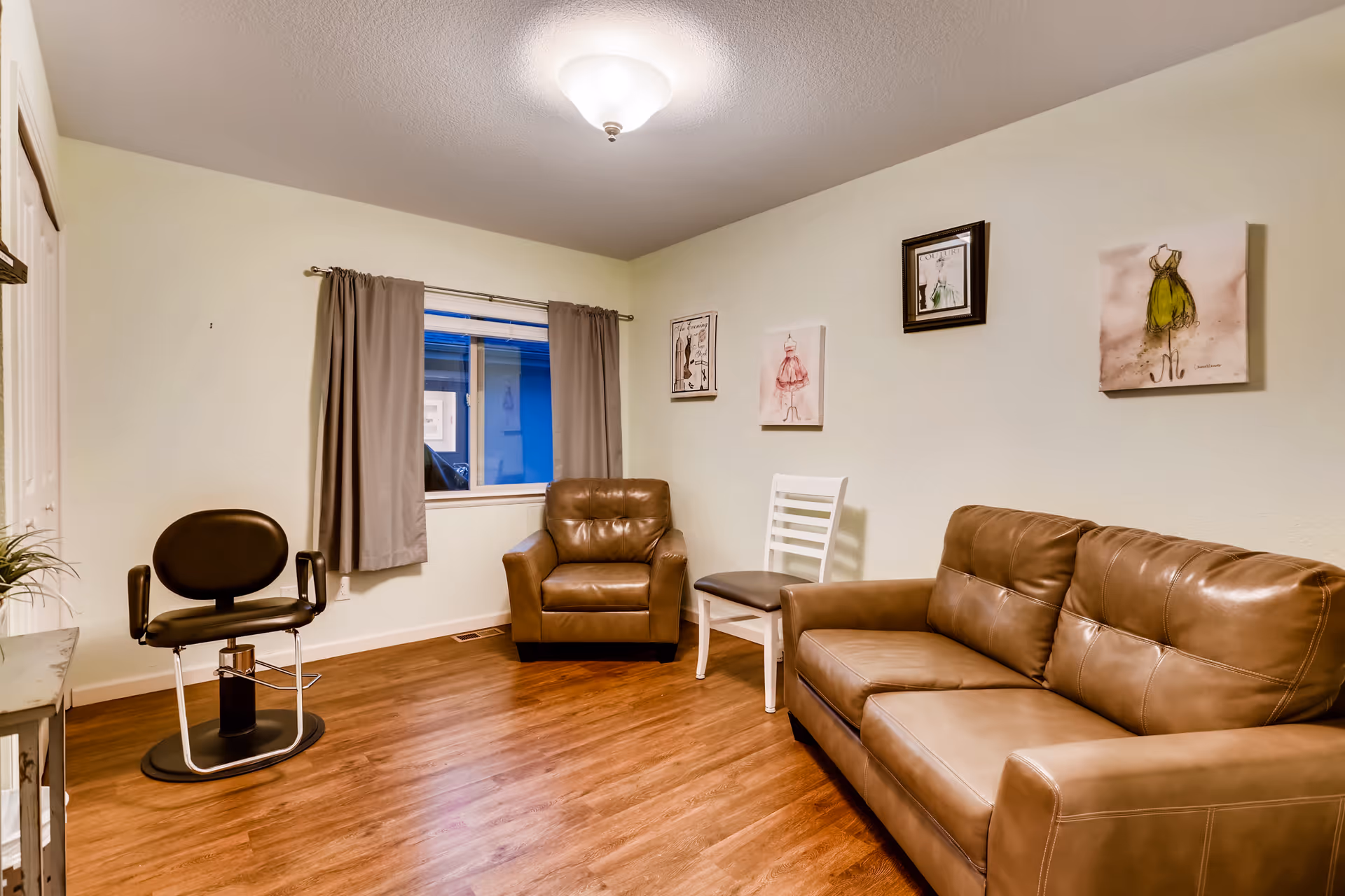 A cozy room with light-colored walls and wooden flooring featuring a brown leather sofa, a matching leather armchair, a white wooden chair with a dark cushion, and a black swivel chair. There is a window with gray curtains and three framed artworks on the wall. The room is lit by a ceiling light fixture.