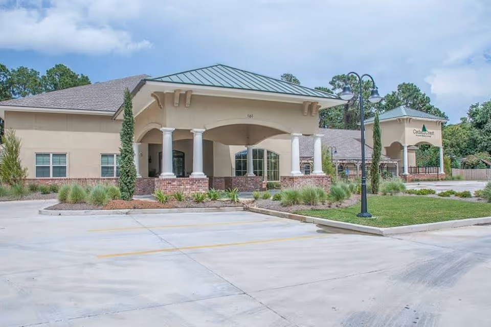 Exterior view of Cedar Crest Memory Care facility showing a single-story building with a covered entrance supported by white columns, surrounded by landscaped greenery and a parking area in front.