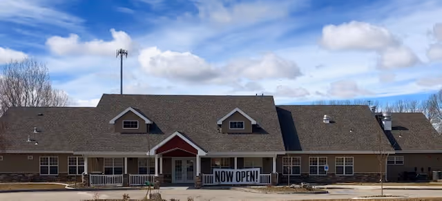 Single-story assisted living building with a covered front porch and a 'NOW OPEN!!' banner under a partly cloudy sky.